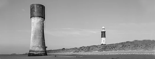 Black and white landscape of two coastal lighthouses, one tall and weathered, one smaller, standing near rshoreline at Spurn Point