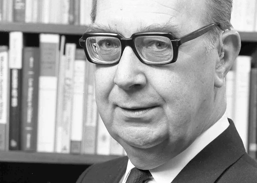 Black and white portrait of Philip Larkin with glasses, wearing a suit, standing in front of a bookshelf