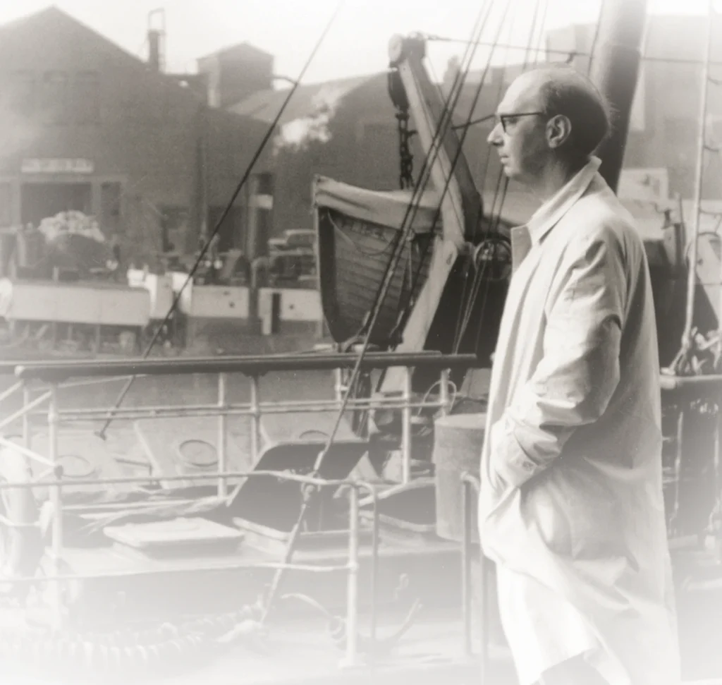 Black and white photo of Larkin in glasses standing on a ship deck, industrial background with boats and buildings