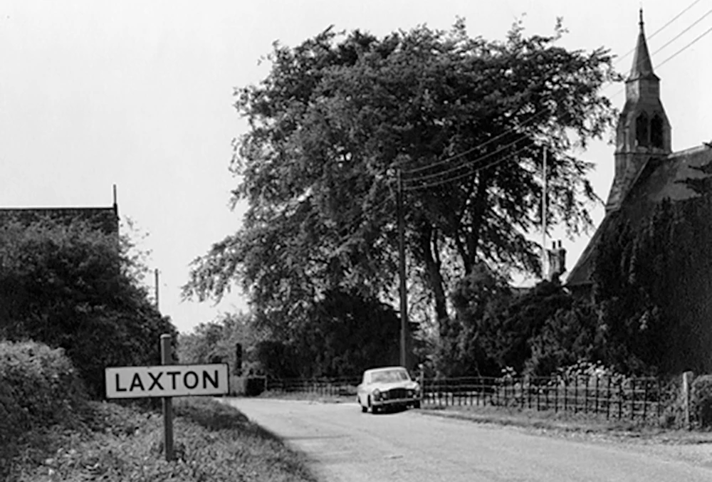 Black and white photo of Laxton village sign, vintage car, church steeple, and tree-lined road.