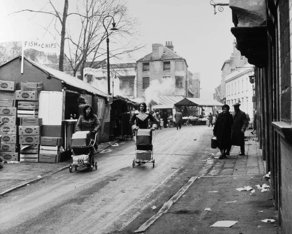 Black and white street scene of Hull market Place with fish and chips stalls, people pushing carts, vintage urban setting