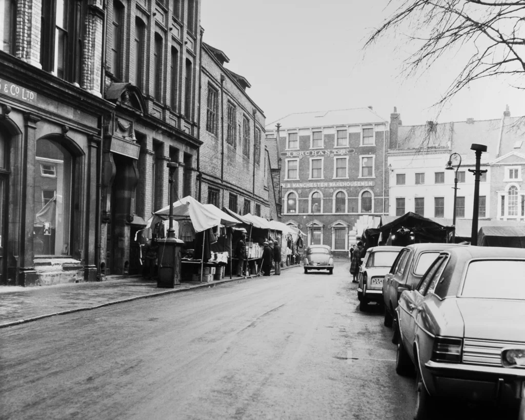 Black and white photo of market stalls, vintage cars, and historic brick buildings in urban setting